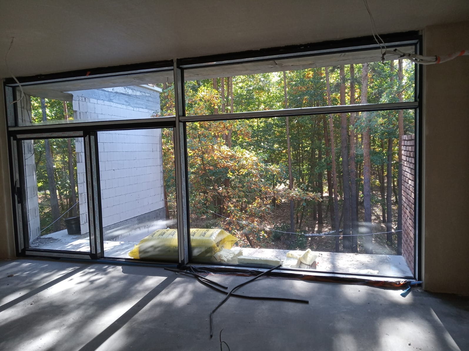 Duże, panoramiczne okno z czarnymi ramami w surowym wnętrzu. Widok na las zza okna. Częściowo wykończony budynek w tle. Elementy montażowe na podłodze.