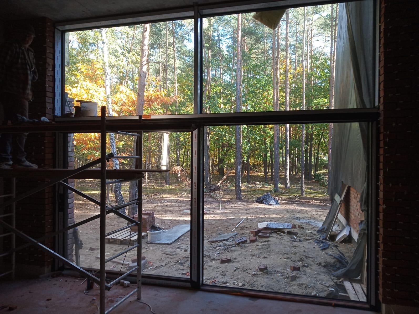 Montaż okna aluminiowego z widokiem na las jesienią. Widoczne rusztowanie i murarz. Duże przeszklenie w surowym stanie budynku.