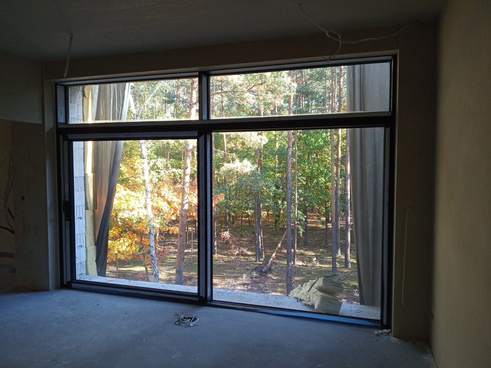 Duże, panoramiczne okno z czarną ramą, widok na jesienny las. Wnętrze w stanie surowym, widoczne materiały izolacyjne. Okno przesuwne, dwuczęściowe.