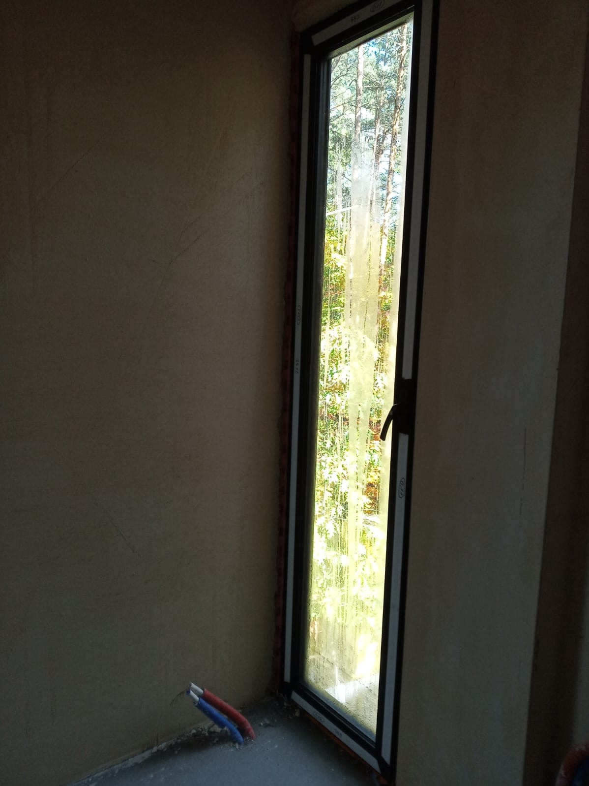 Zamglone okno PCV z widokiem na zieleń w Miedźno, osadzone w surowej ścianie. Widoczne wystające rurki instalacyjne przy podłodze. Ujęcie z bliska, wnętrze w trakcie wykończenia.