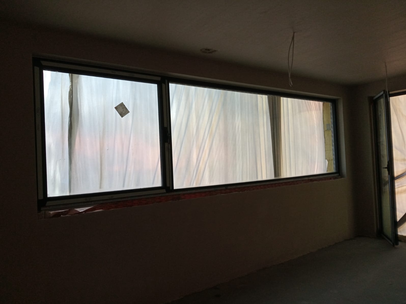 Zamontowane okno aluminiowe z folią ochronną w surowym wnętrzu. Widoczne niedoskonałości wykończenia ściany i brak parapetu. Ujęcie z bliska, wnętrze w trakcie prac wykończeniowych.