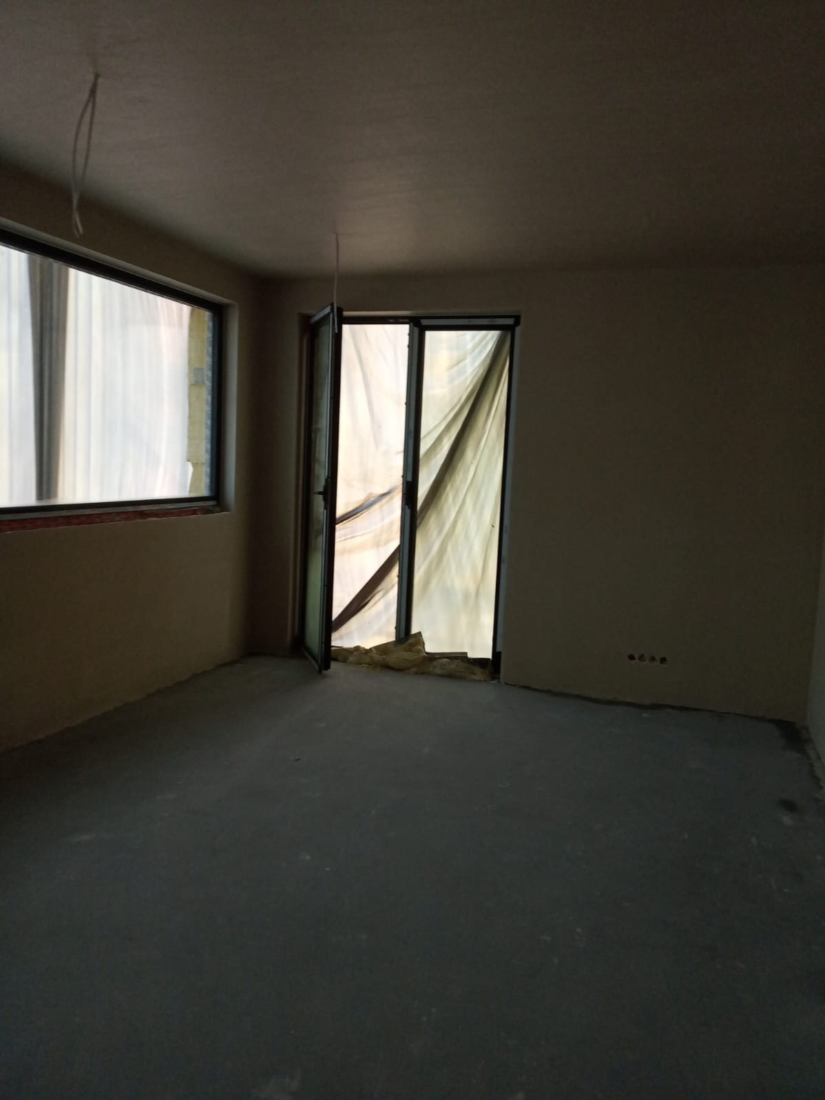 Wnętrze w trakcie wykańczania: okno PCV z czarną ramą, otwarte drzwi balkonowe zabezpieczone folią malarską, surowa podłoga i ściany przygotowane do malowania.