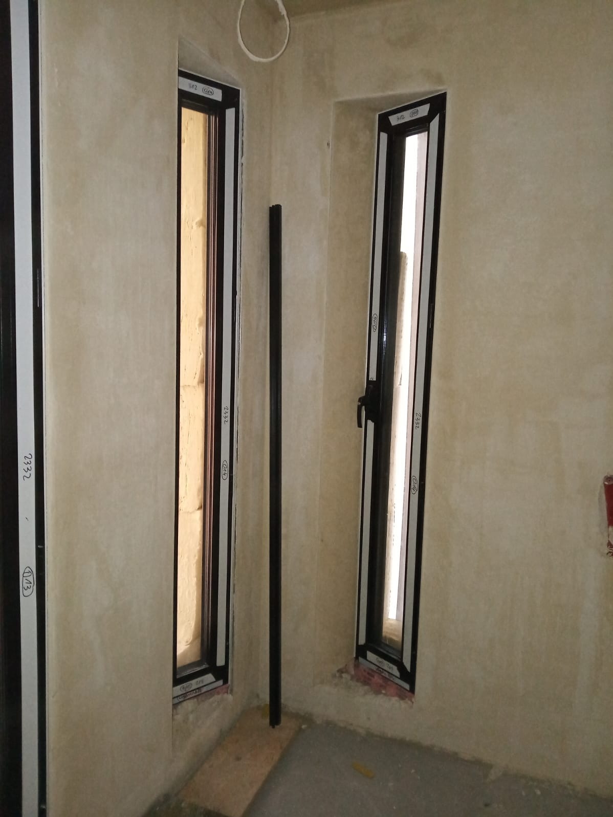 Wnętrze z dwoma wąskimi oknami PCV z czarnymi ramami, zamontowanymi w surowych ścianach. Widoczny profil okienny z naklejkami producenta. Jeden profil oparty o ścianę.