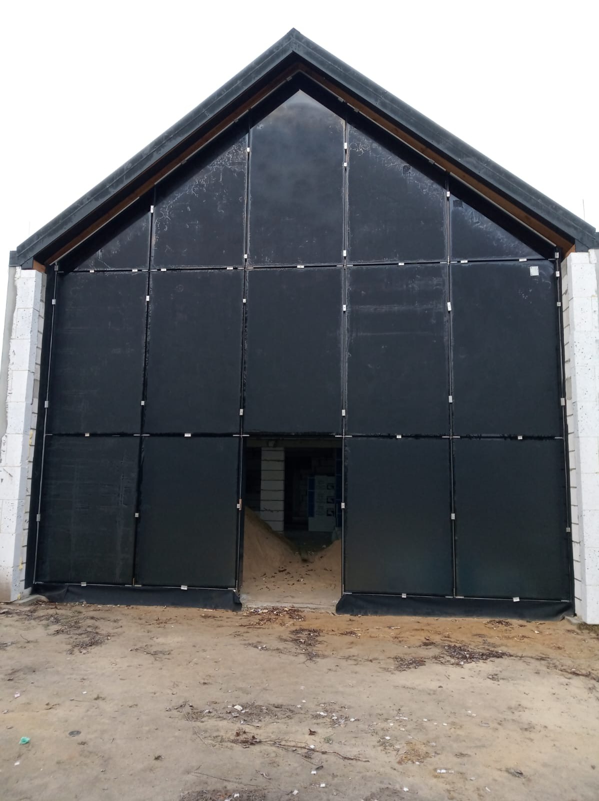 Ciemna fasada hali w trakcie budowy, podzielona na segmenty z widocznymi mocowaniami. Surowy stan, białe bloczki po bokach, ziemiste podłoże.