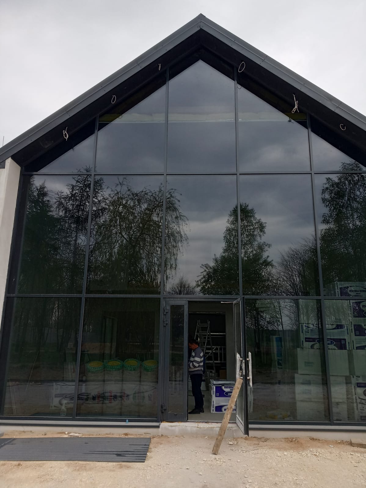Duże przeszklenie budynku z ciemnymi ramami, odbijające drzewa i niebo. W otwartych drzwiach stoi osoba, w tle widoczna drabina i materiały budowlane.