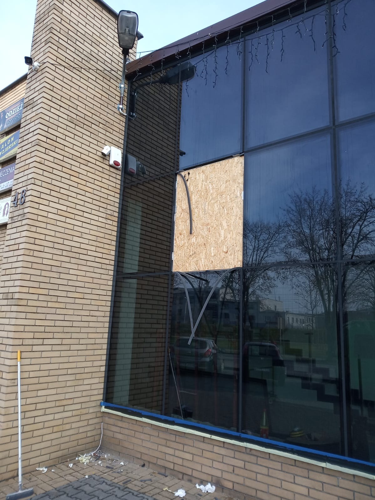 Uszkodzone okno aluminiowe w budynku z cegły, zabezpieczone płytą OSB z wystającą rurką. Widoczne ślady prac naprawczych i odbicie otoczenia w szybach.