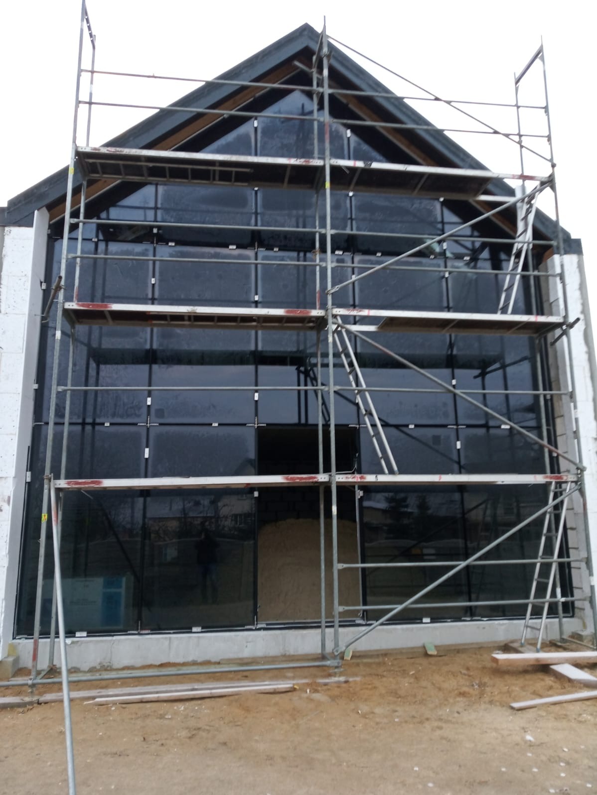 Montaż okien w budynku o nowoczesnej architekturze. Widoczne rusztowania i duże przeszklenia. Budynek w trakcie wykończenia, surowy stan.