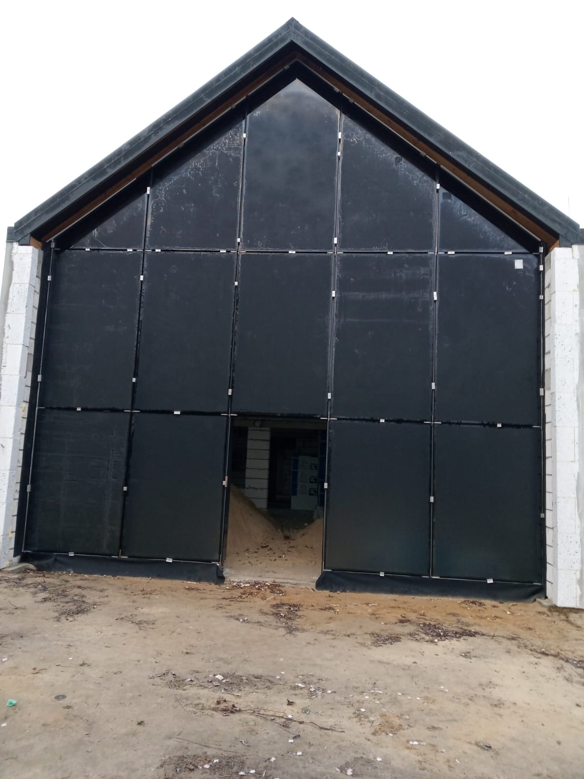 Ciemna fasada budynku w trakcie budowy, podzielona na panele z widocznymi mocowaniami. Surowy stan, widoczne bloczki i ziemia. Ujęcie z dołu, podkreślające wysokość.