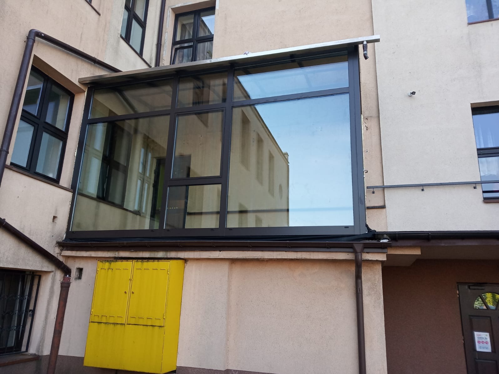 Zabudowa balkonu z ciemną ramą okienną, widoczna żółta skrzynka gazowa na ścianie budynku. W szybach odbija się sąsiedni budynek.