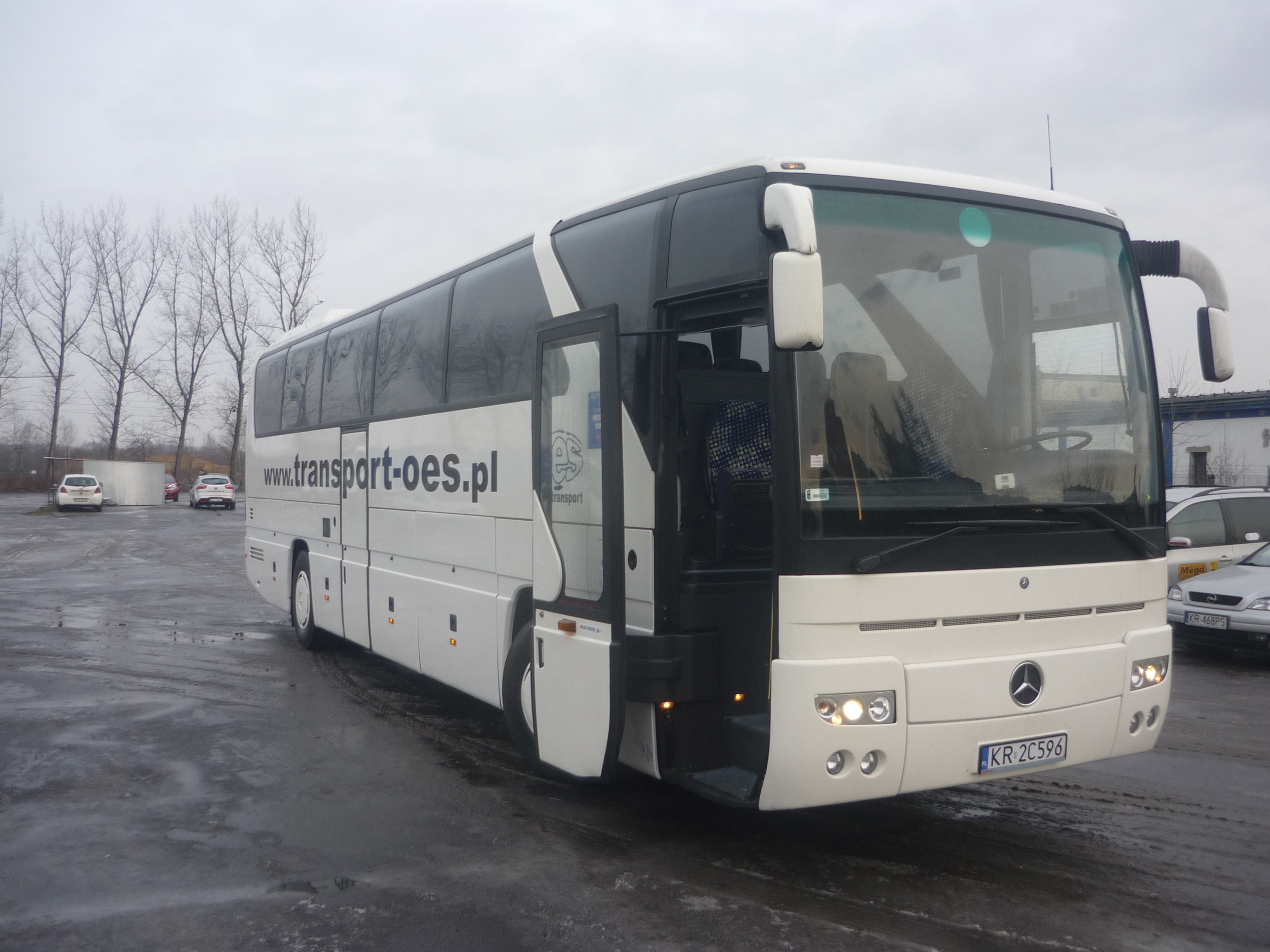 Biały autokar z otwartymi drzwiami na mokrym parkingu, widoczny numer rejestracyjny KR 2C596 i adres strony internetowej transport-oes.pl.