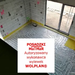 Przygotowanie do wylewki z ogrzewaniem podłogowym w surowym wnętrzu, widoczne rury grzewcze ułożone na folii z siatką, ściany z betonu komórkowego z oznaczeniami, okno z widokiem na zewnątrz.