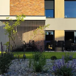 Nowoczesny ogród w Dominowie z rabatą wysypaną grysem, młodym drzewkiem i kwitnącymi bylinami na tle budynku z ceglaną elewacją i ażurową przesłoną tarasu.