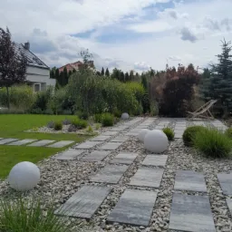 Nowoczesny ogród z ścieżką z szarych płyt ułożonych na żwirze, ozdobne kule, trawa ozdobna, krzewy i drzewa w tle, widok na dom.