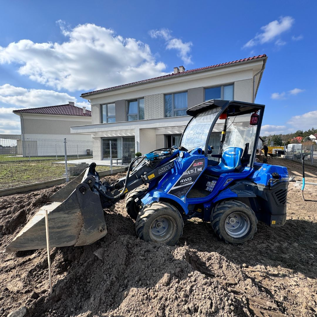 Niebieska mini ładowarka na pryzmie ziemi przed nowoczesnym domem jednorodzinnym z białej cegły, błękitne niebo w tle.