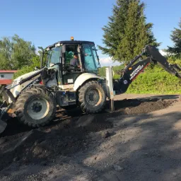 Biało-czarna koparko-ładowarka marki Hydromek podczas prac ziemnych, widoczny operator w kabinie, świeżo naruszona warstwa ziemi i drzewa w tle.