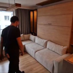 MEB-MAJSTER DAWID BIESZKE - Jasna sofa narożna z funkcją spania, wkomponowana w zabudowę meblową z drewnianymi półkami. Widok na okno z dekoracją w kształcie gwiazdy i fragment kuchni.