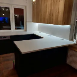 Nowoczesna kuchnia z czarnymi szafkami i jasnym blatem roboczym, oświetlona listwą LED pod szafkami wiszącymi z drewnianym frontem. Widoczne gniazdka w blacie.