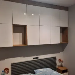 Białe szafki wiszące nad łóżkiem z drewnianymi wnękami po bokach, oświetlone lampkami nocnymi.