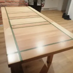 Rozkładany stół z jasnego drewna z zielonymi intarsjami, widoczny w domowym wnętrzu z ceglaną ścianą w tle.