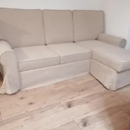 Jasna, trzyosobowa kanapa z szezlongiem po prawej stronie, obita beżową tkaniną z delikatnym obramowaniem, ustawiona na drewnianej podłodze.