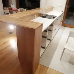 Jasna kuchnia z drewnianym blatem i półwyspem, płyta grzewcza wkomponowana w blat, białe szafki z czarnymi uchwytami, drewniana podłoga i białe krzesło w tle.