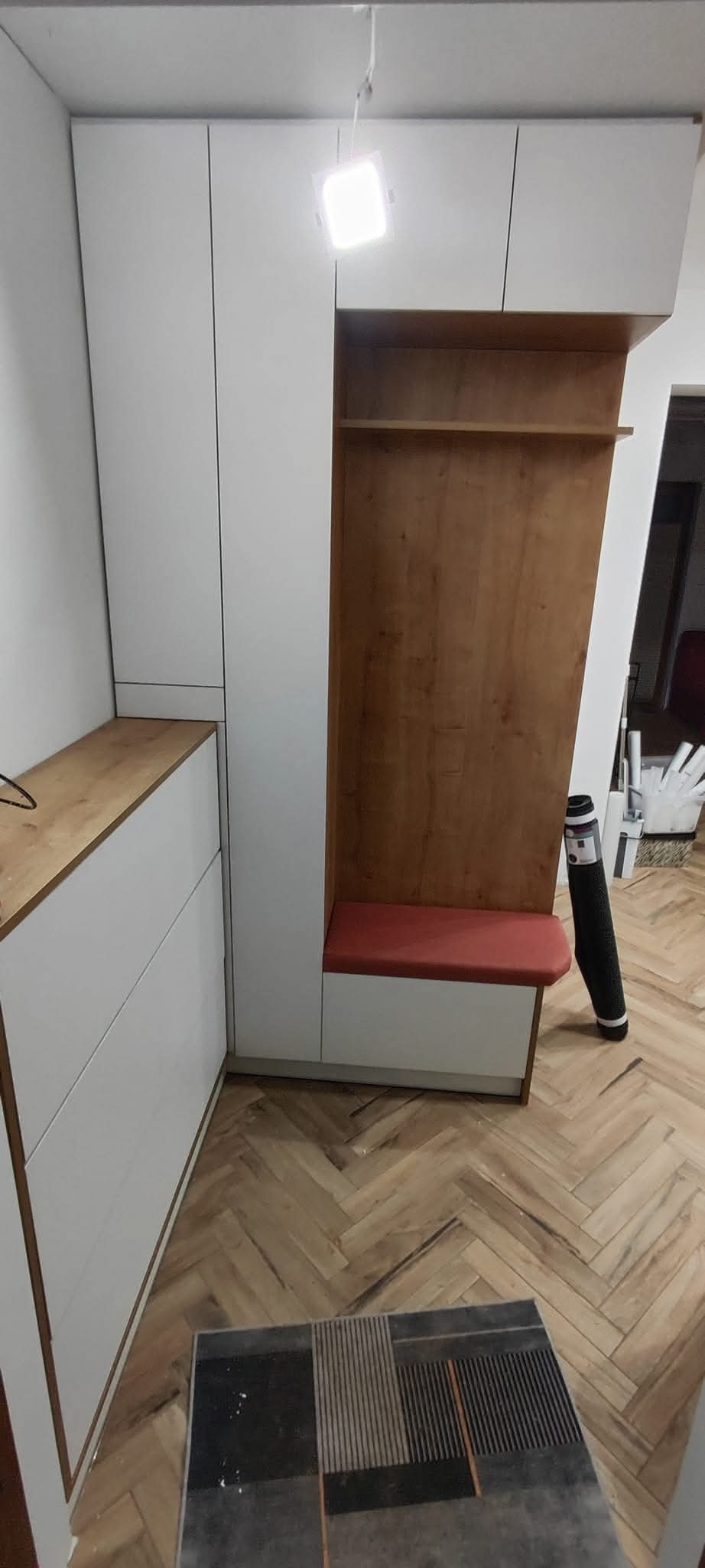 Biała szafa na wymiar z drewnianym blatem i siedziskiem z czerwoną tapicerką, w przedpokoju z parkietem ułożonym w jodełkę. Nowoczesny design.