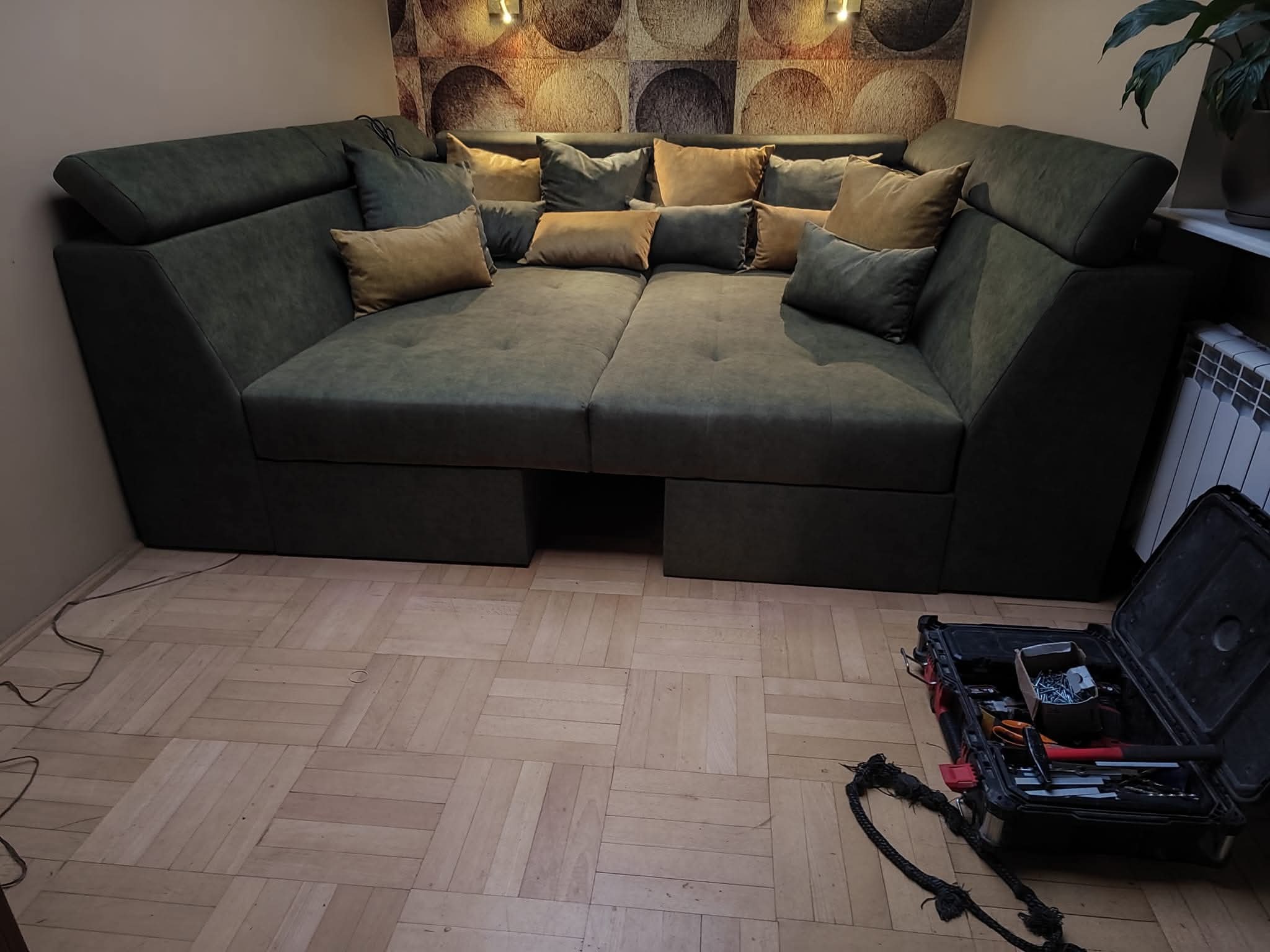 Rozłożysty, zielony narożnik z poduszkami w salonie, na podłodze parkiet. W rogu widoczna skrzynka z narzędziami, sugerująca montaż lub naprawę mebla.