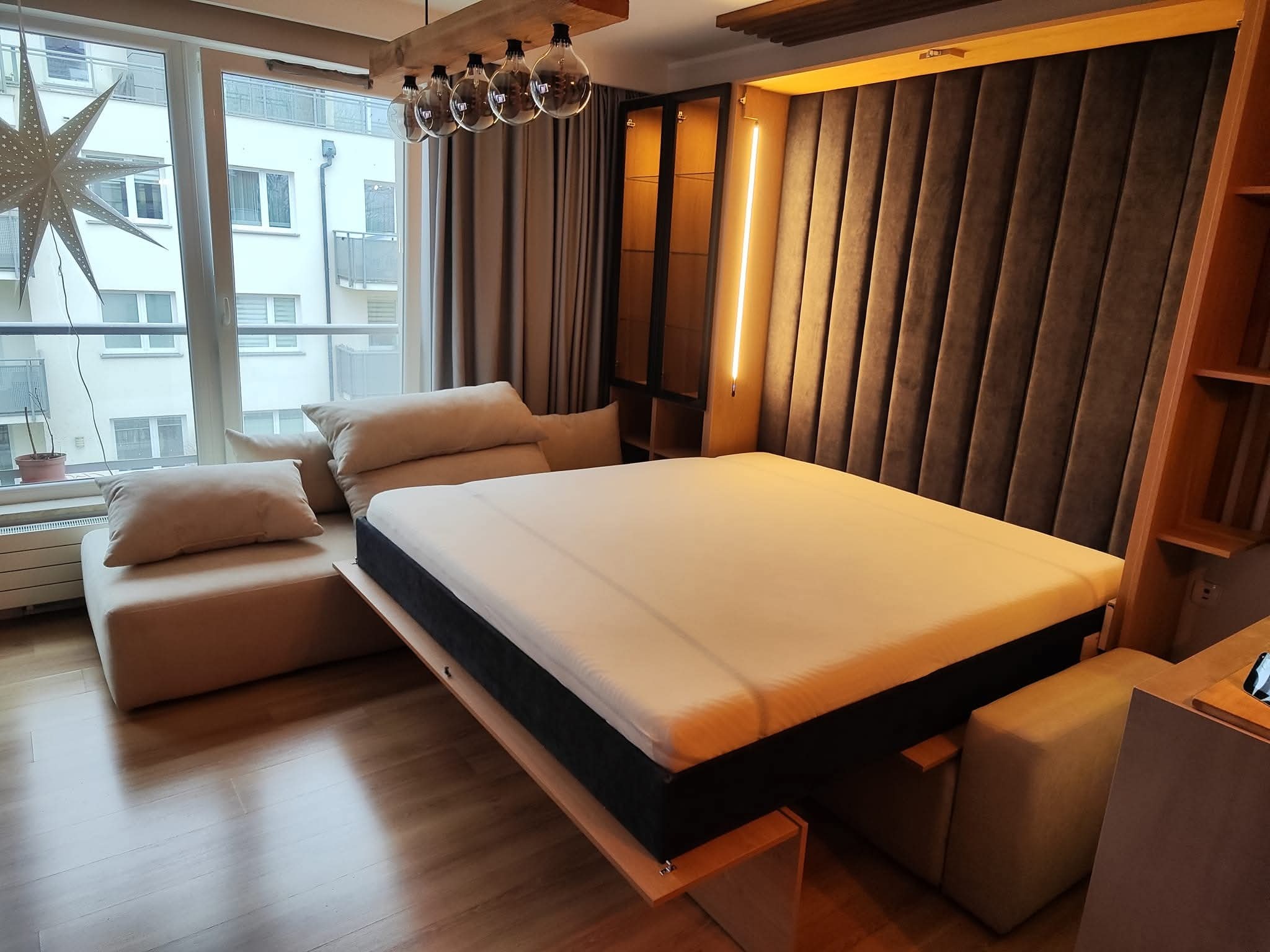 Rozłożone łóżko w szafie z tapicerowanym wezgłowiem i oświetleniem LED, widok na okno z dekoracyjną gwiazdą. Nowoczesny design, jasne wnętrze.