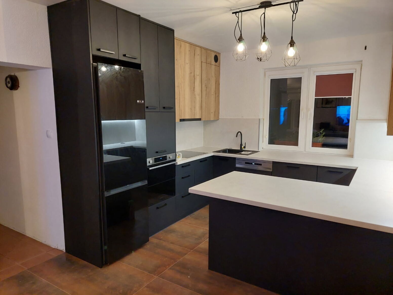 Nowoczesna kuchnia z czarnymi szafkami, drewnianymi akcentami i wyspą z białym blatem. Oświetlenie punktowe nad wyspą. Widok na okno z roletą.