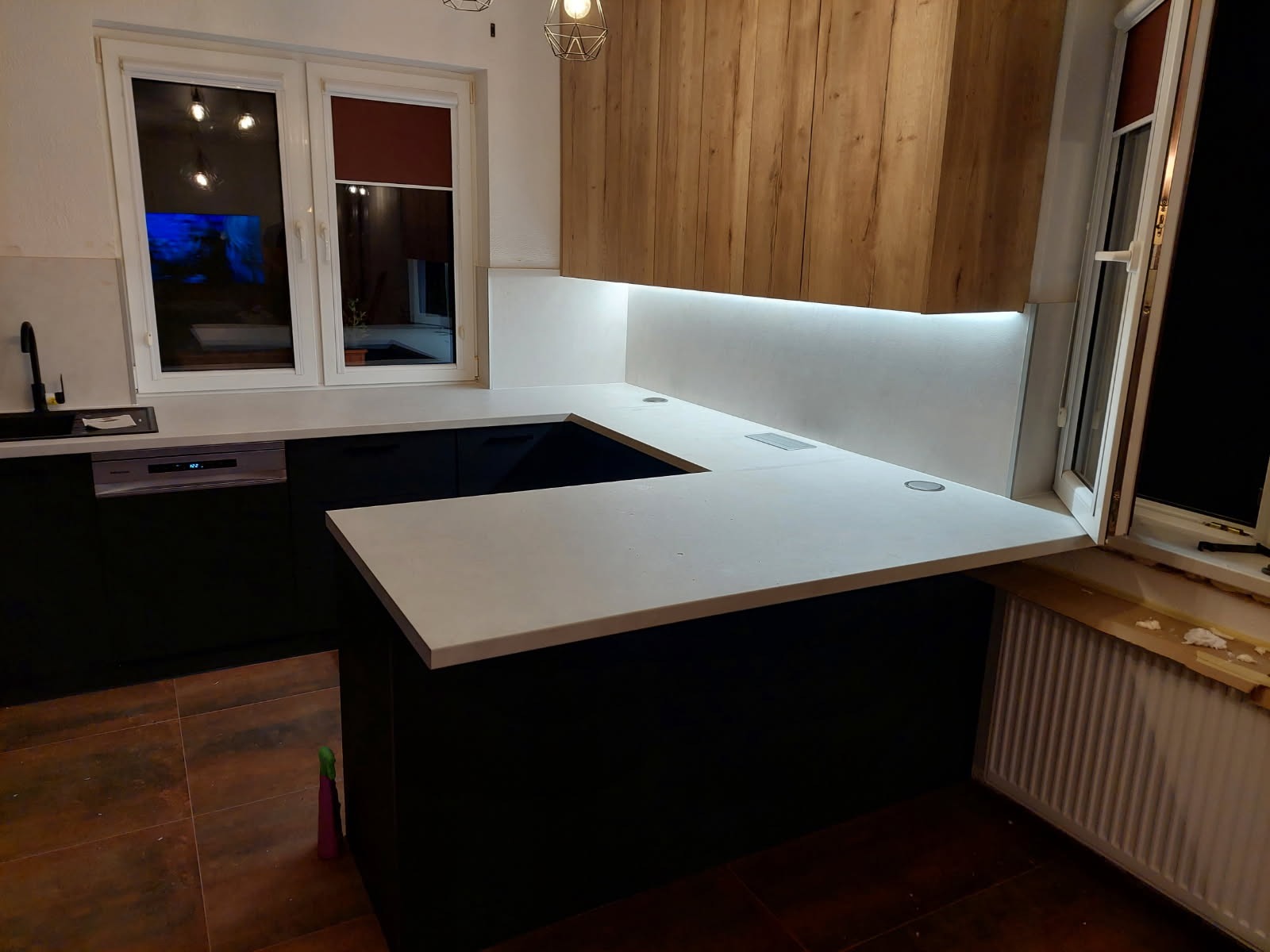 Nowoczesna kuchnia z czarnymi szafkami i jasnym blatem roboczym, oświetlona listwą LED pod szafkami wiszącymi z drewnianym frontem. Widoczne gniazdka w blacie.