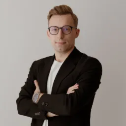 Portret mężczyzny w okularach i czarnej marynarce, z założonymi rękami i widocznym zegarkiem na nadgarstku.