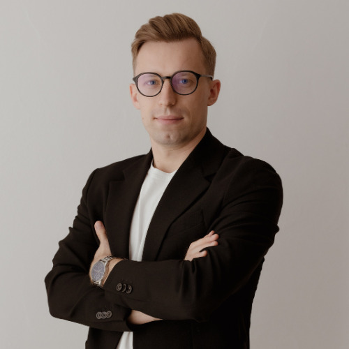 Portret mężczyzny w okularach i czarnej marynarce, z założonymi rękami i widocznym zegarkiem na nadgarstku.