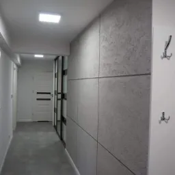 Nowoczesny korytarz z szarą ścianą wykończoną panelami imitującymi beton, oświetlenie punktowe na suficie i minimalistyczne wieszaki na ścianie.