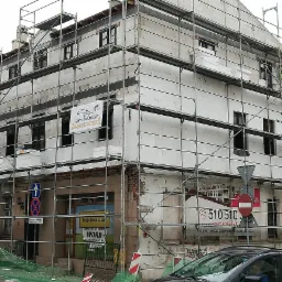 Budynek w trakcie docieplania styropianem, widoczne rusztowania, pracownik w kamizelce odblaskowej na rusztowaniu, baner reklamowy na elewacji, znaki drogowe w tle.