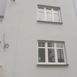 Jasnoszara elewacja budynku z dwoma rzędami białych okien, widoczne przewody elektryczne na ścianie.