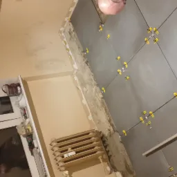 Szare płytki podłogowe układane z użyciem żółtych krzyżyków dystansowych w trakcie remontu łazienki, widoczny grzejnik i okno.