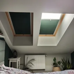Dwa okna dachowe w sypialni na poddaszu. Jedno okno z całkowicie zaciemniającą plisą w kolorze czarnym, drugie z plisą w kolorze zielonym. W tle widoczny grzejnik i rośliny doniczkowe.