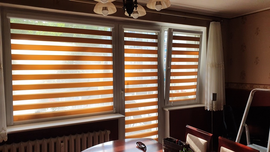 Pomarańczowo-białe rolety dzień-noc w kasecie z prowadnicami PCV, zamontowane w trzech oknach, z widocznym fragmentem pokoju z brązowymi ścianami i zasłonami.
