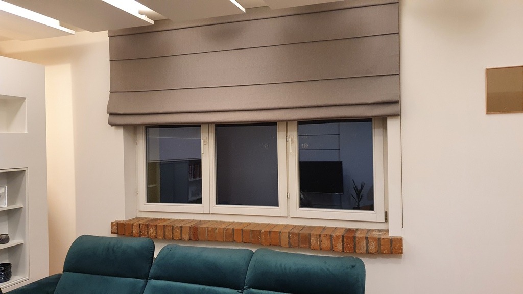 Zamknięta, szara, materiałowa osłona okienna z poziomymi zakładkami, zamontowana nad trzema oknami z ceglanym parapetem w jasnym wnętrzu z fragmentem zielonej sofy.