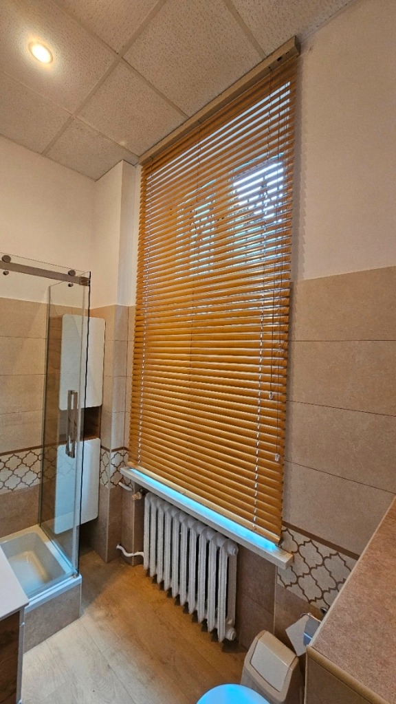 Jasne, poziome lamele w oknie łazienki, widoczne przy grzejniku żeberkowym i kabinie prysznicowej z przezroczystymi drzwiami.