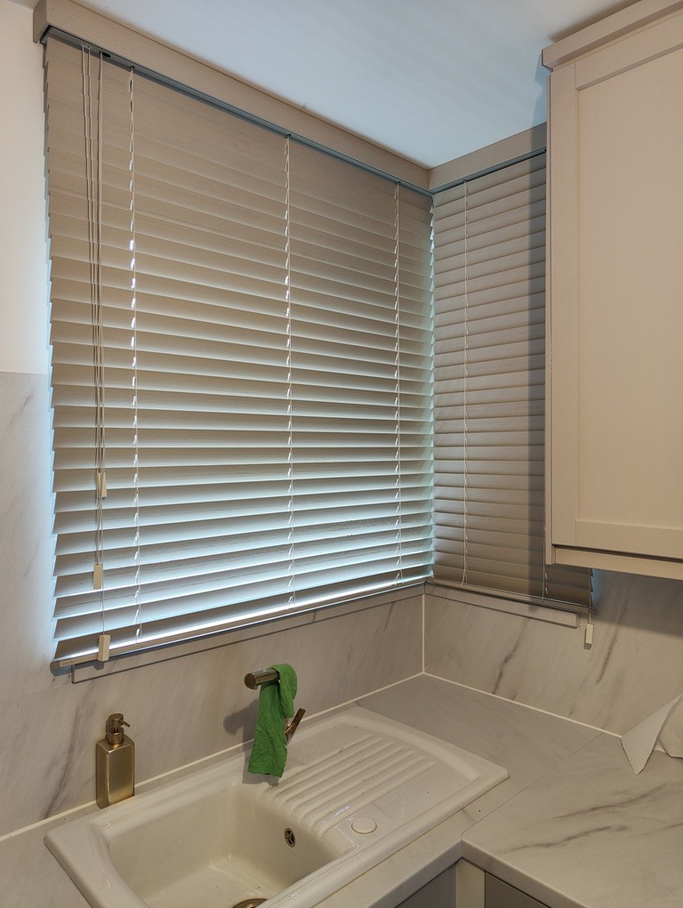 Jasne, poziome listwy osłaniają okno nad zlewem w kuchni; widoczne sznurki sterujące i fragment szafki kuchennej.