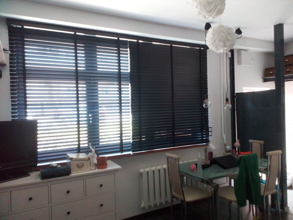 Ciemne poziome osłony okienne z drewna, zamontowane w oknach w jasnym pokoju dziennym z białymi ścianami, widoczne z perspektywy wnętrza, z fragmentem komody i stołu.