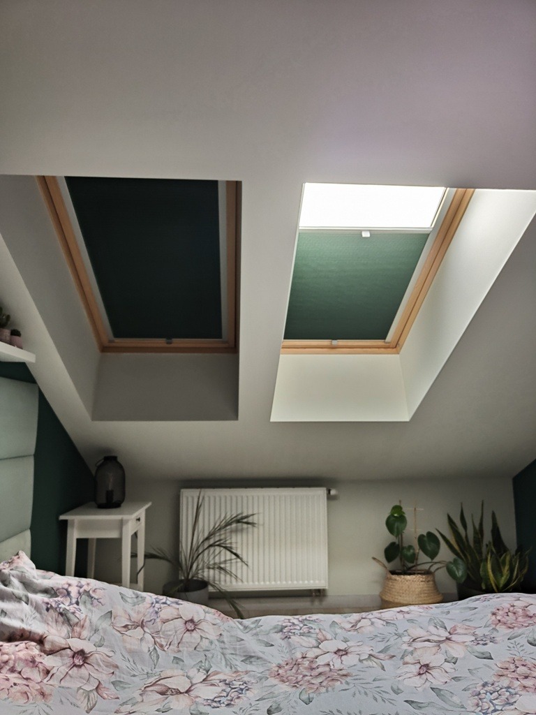 Dwa okna dachowe w sypialni na poddaszu. Jedno okno z całkowicie zaciemniającą plisą w kolorze czarnym, drugie z plisą w kolorze zielonym. W tle widoczny grzejnik i rośliny doniczkowe.