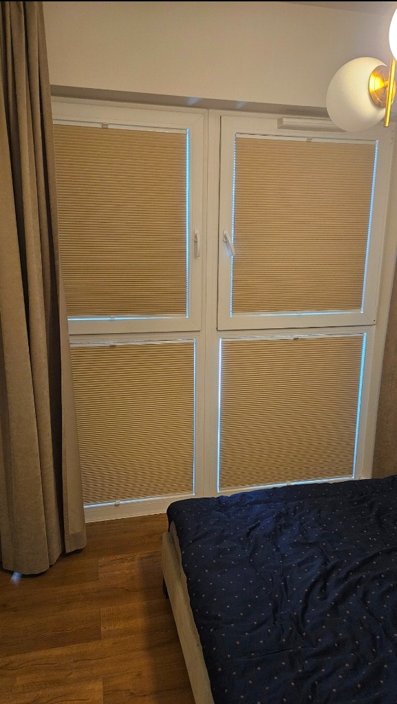 Beżowe, poziome przesłony okienne typu plaster miodu zamontowane w czterech segmentach okna, widoczne z wnętrza sypialni z fragmentem łóżka i zasłony.