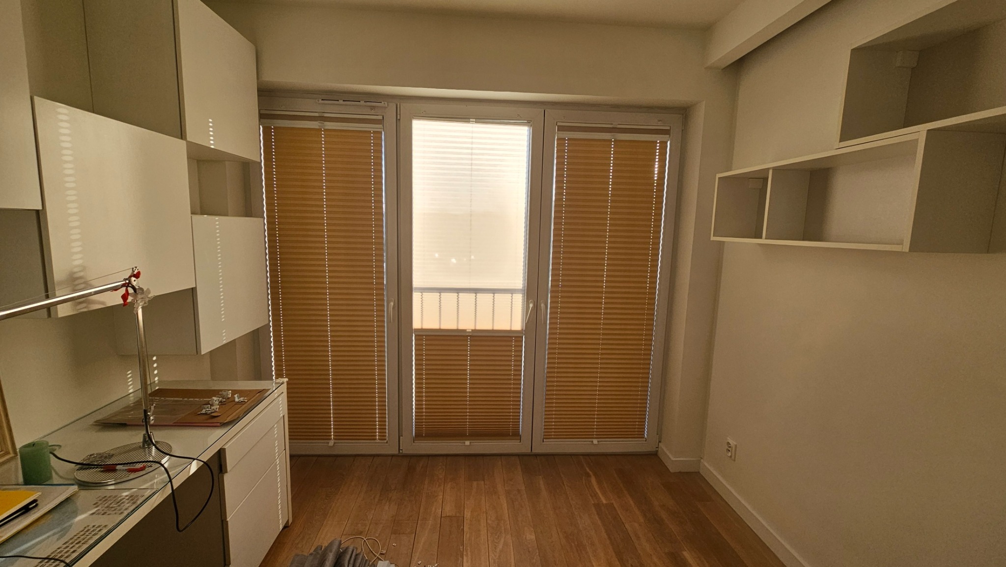 Beżowe poziome osłony okienne zamontowane w oknach balkonowych w pokoju z drewnianą podłogą i białymi meblami.