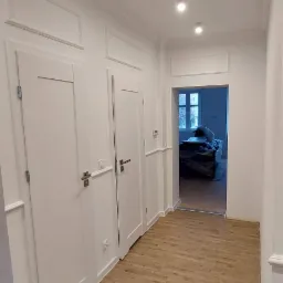 Korytarz w jasnych barwach po remoncie, z drewnopodobnymi płytkami na podłodze ułożonymi w jodełkę, białymi drzwiami z frezowanymi panelami i dekoracyjnymi listwami przysufitowymi.