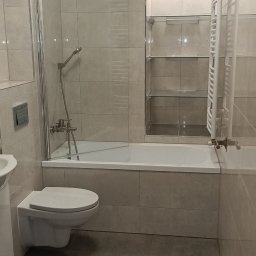 RC usługi ogólnobudowlane - Jasna łazienka po remoncie: beżowe płytki, wanna z parawanem, podwieszana toaleta i grzejnik drabinkowy. Minimalistyczny design z elementami chromu.