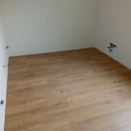 Jasne panele podłogowe ułożone w rogu pomieszczenia z białymi ścianami, widoczne puszki elektryczne i fragment ościeżnicy.