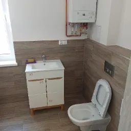 Wykończona łazienka z brązowymi płytkami imitującymi drewno, białą szafką pod umywalką z fioletową gąbką, wiszącą toaletą z otwartą klapą oraz białym piecem gazowym na ścianie.