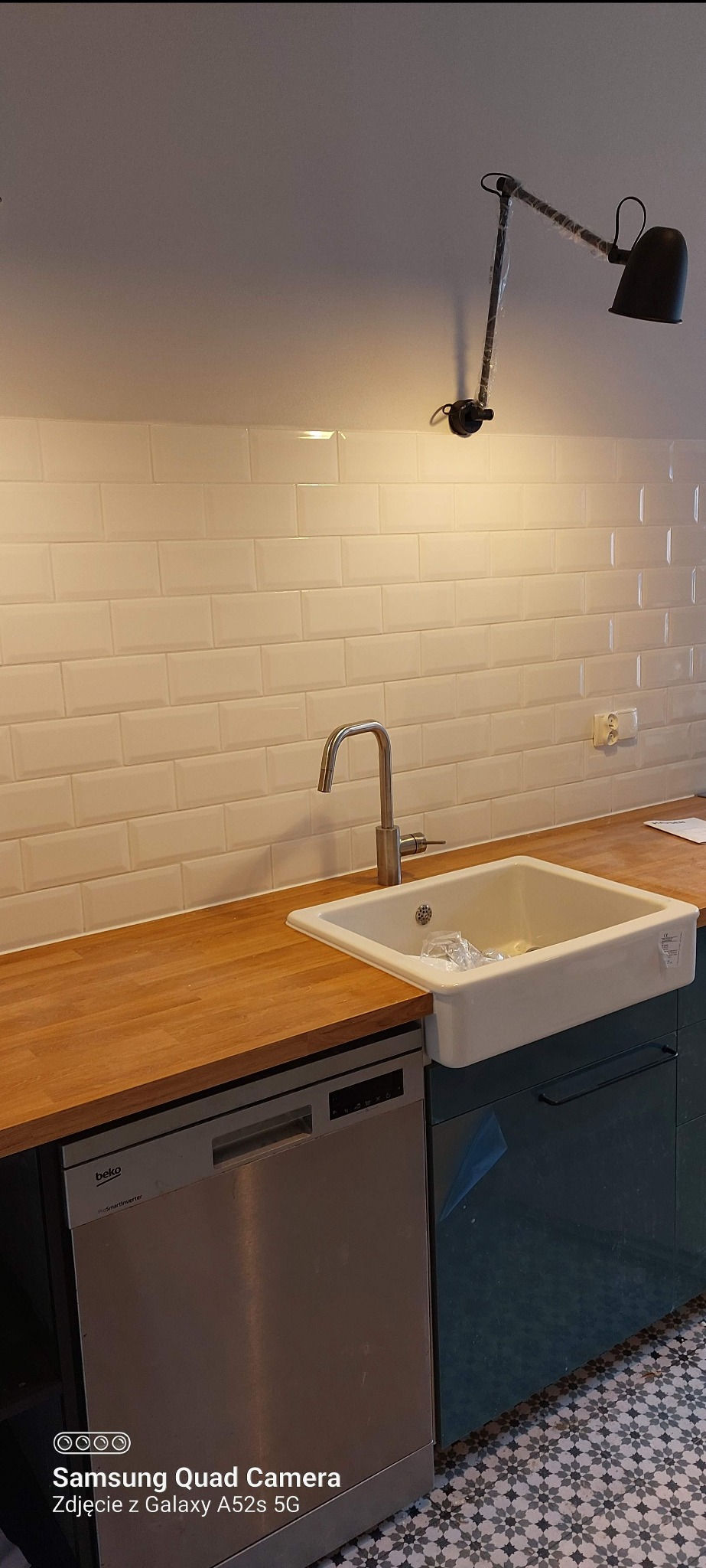 Nowoczesna kuchnia z białymi płytkami metra, drewnianym blatem, ceramicznym zlewem w stylu farmhouse, srebrną baterią i zmywarką Beko; lampa z regulowanym ramieniem zamontowana na suficie.